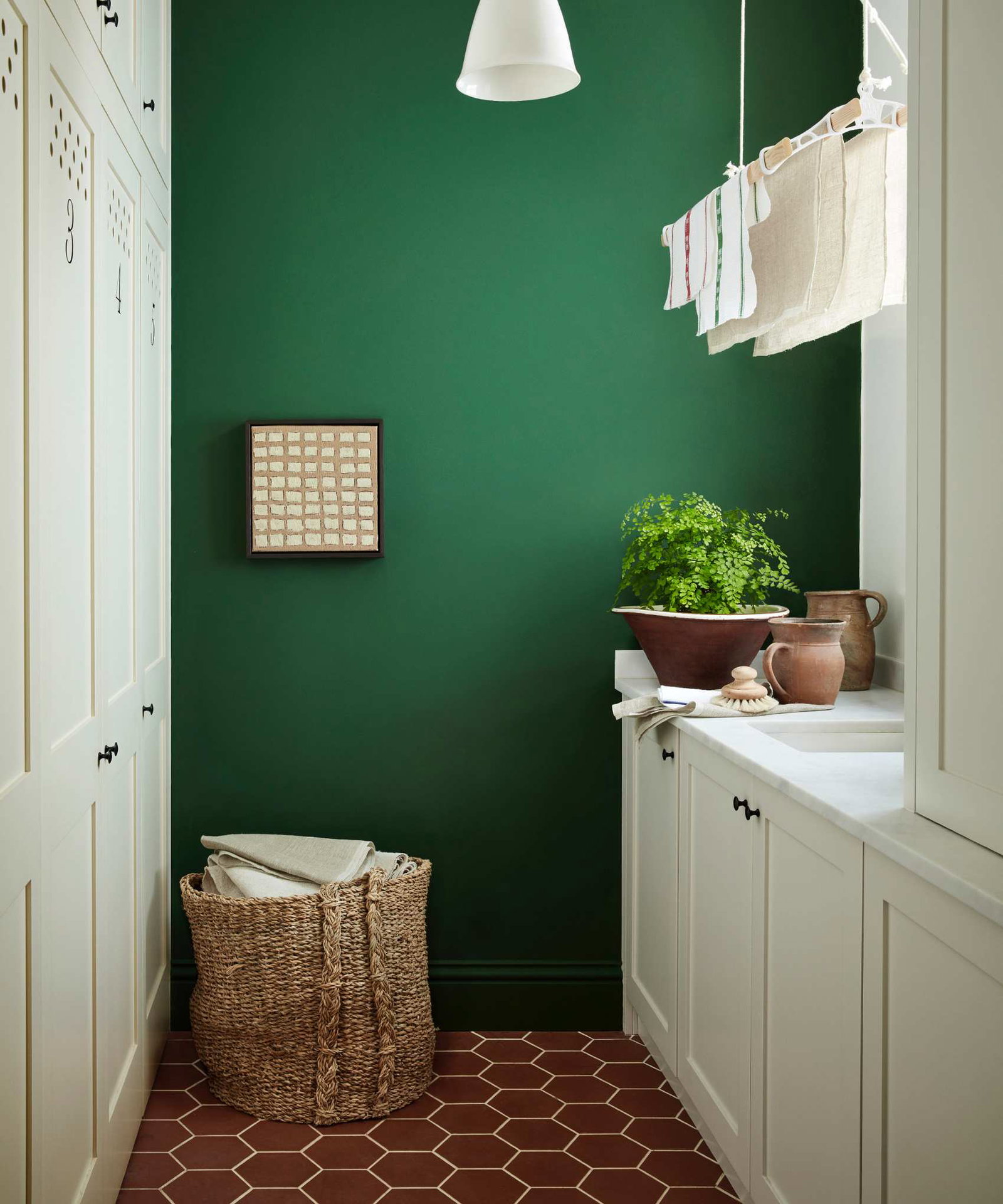 small laundry room with hexagon floor tiles, off white cabinetry, laundry maid, green wall, basket full of linen, sink, pendant light, ceramics
