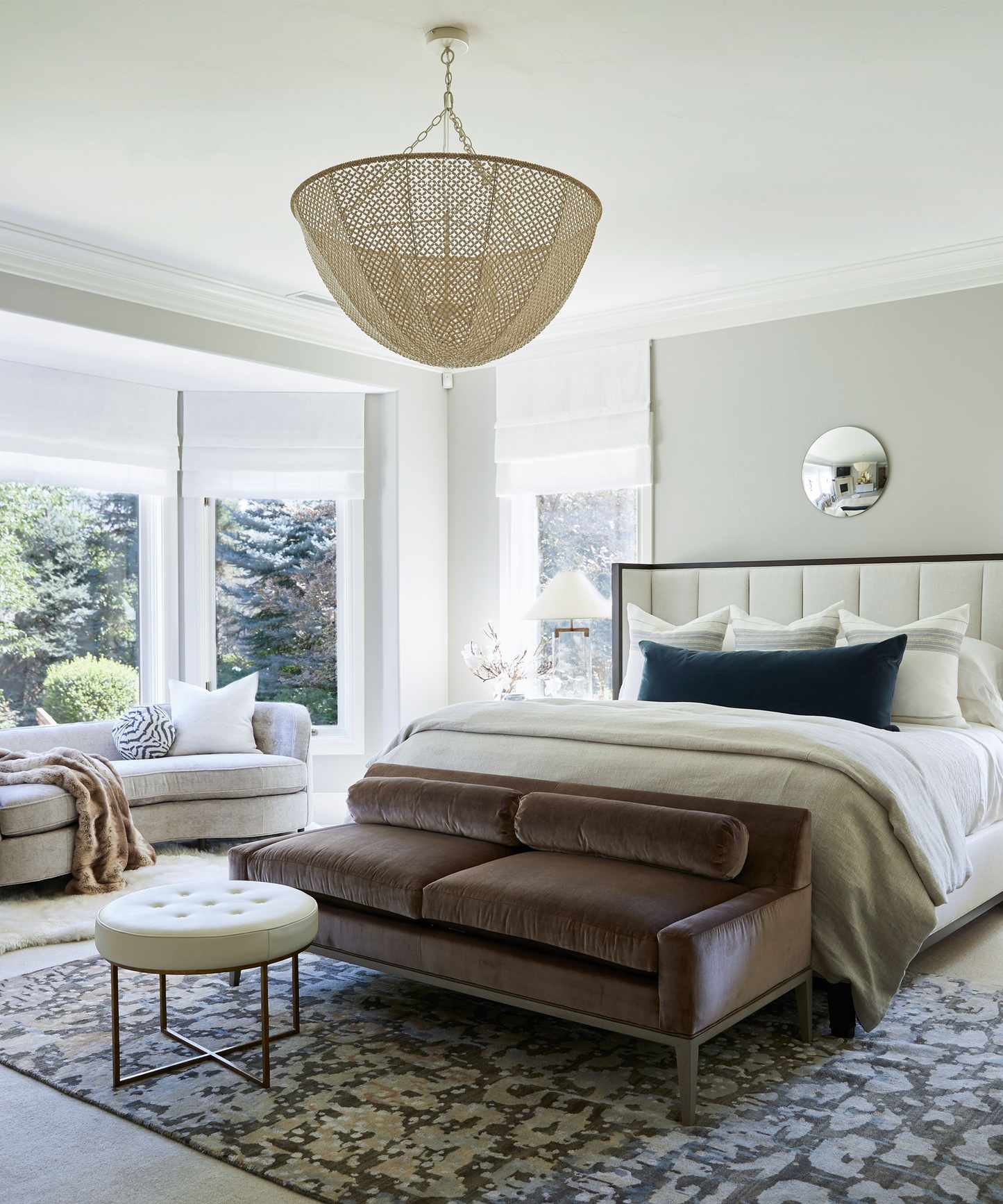 Contemporary, light and bright bedroom with double bed, rounded small mirror mounted on wall above, large textured pendant hanging above bed, brown small sofa at foot of bed, large patterned grey and brown rug at end of bed, sofa in large bay window area
