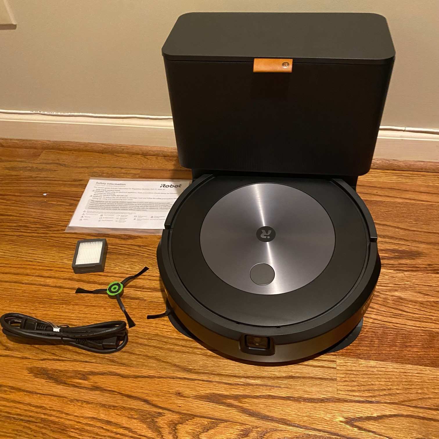The iRobot Roomba j7+ robot vacuum, cleaning base, brush, cable, manual and filter on wooden floor in Jaclyn's home