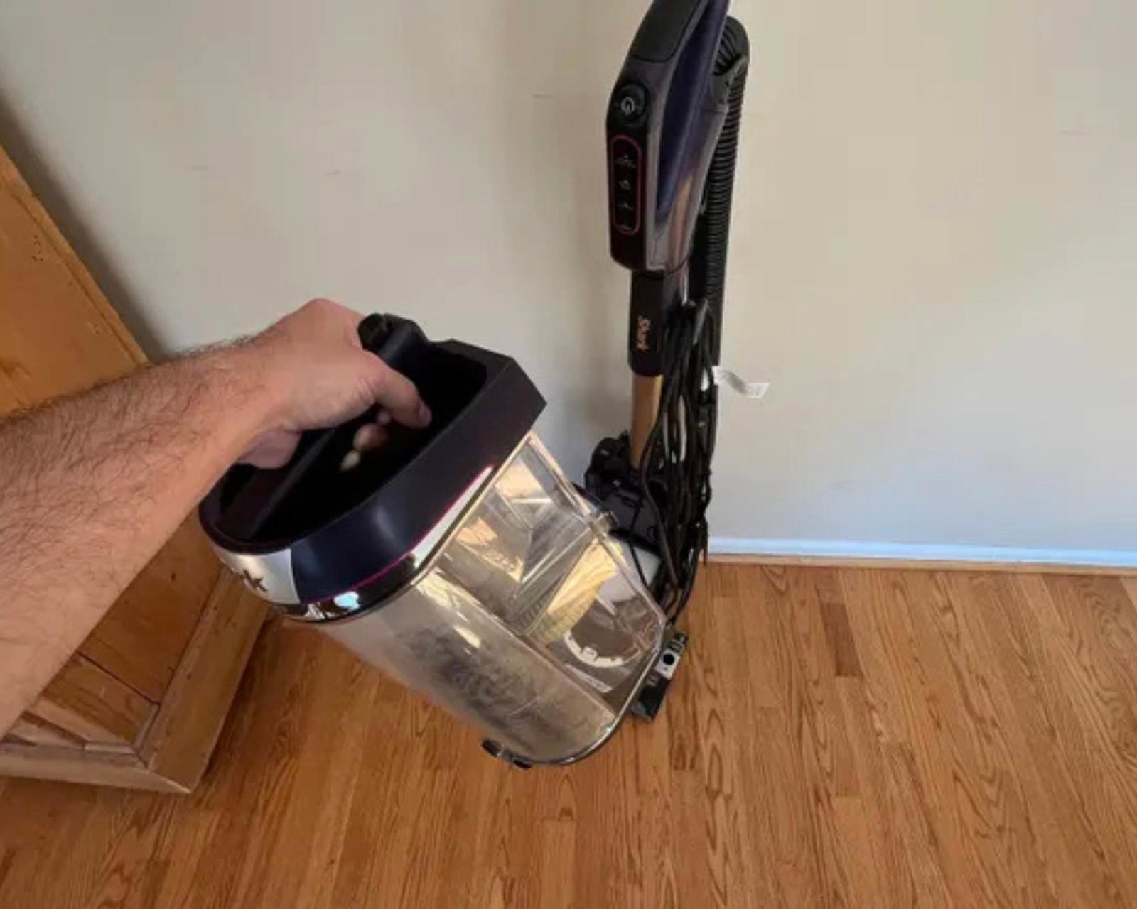 Hand holding the Shark POWERDETECT Upright Vacuum's dust bin  in front of the vacuum on wooden floor.