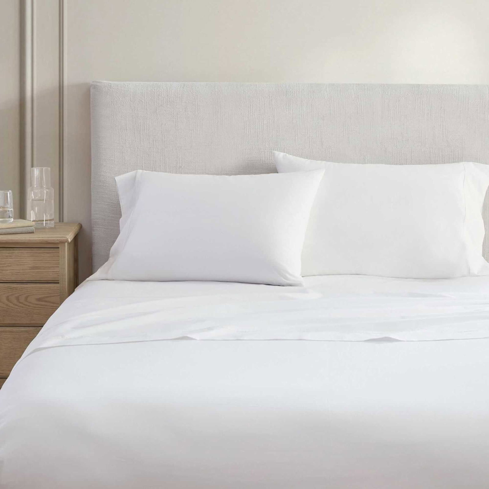 A white Martha Stewart Cotton Percale sheet set pictured on a pale white bed with a white panelled wall behind and wooden beside table with glasses to the left.