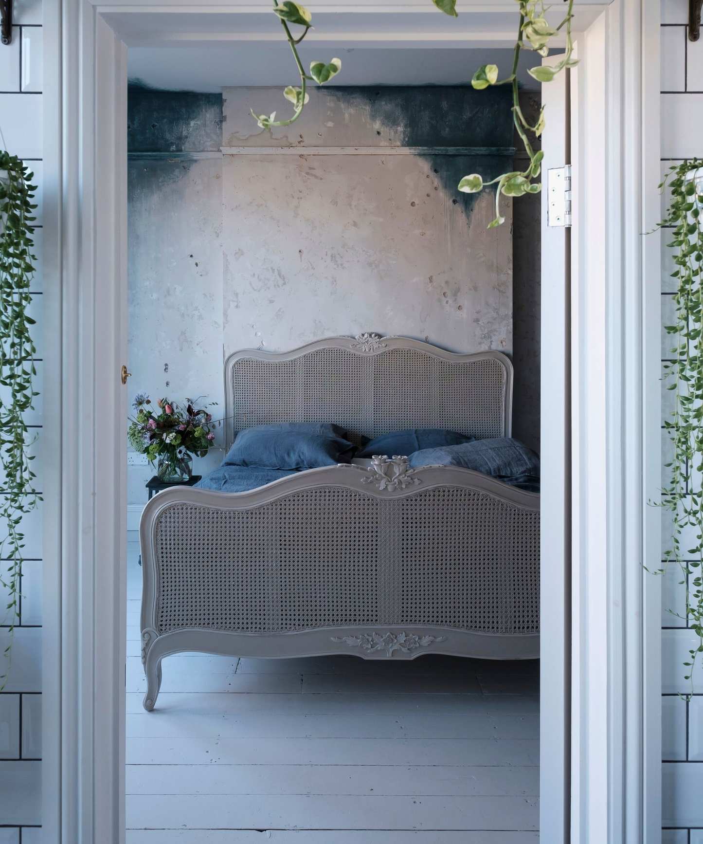View through a doorway from the bathroom into the master bedroom with white wooden floorboards, blue stained walls, and French cane bed.